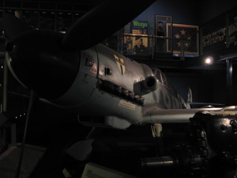 1991 Ford LTD Crown Victoria - nose, spinner, and propeller of a gray German Messerschmitt fighter with shield emblem on display in a dim museum hall.