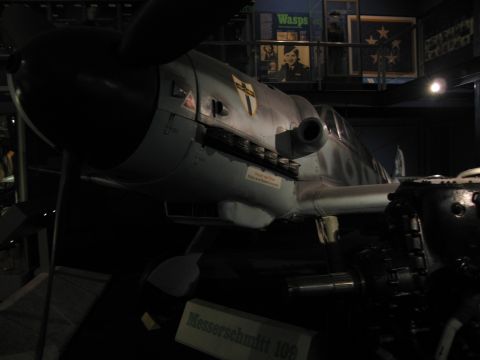 1991 Ford LTD Crown Victoria - gray nose and fuselage of a Messerschmitt 109 fighter with shield emblem displayed above a labeled museum sign in a dark hall.