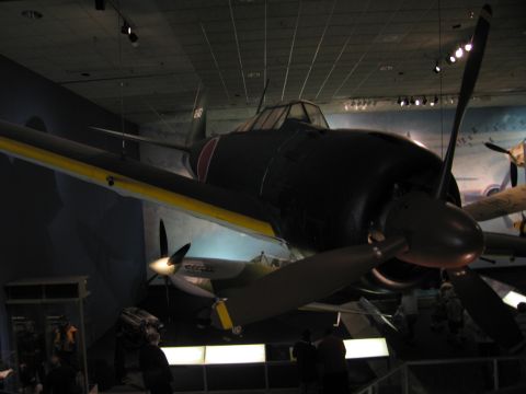1991 Ford LTD Crown Victoria - dark WWII single-engine fighter with yellow wing-leading-edge bands, mounted high above visitors on a museum floor.