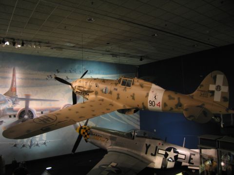 1991 Ford LTD Crown Victoria - desert-camouflage WWII fighter marked 90-4 suspended in a museum above another aircraft bearing a US star insignia.