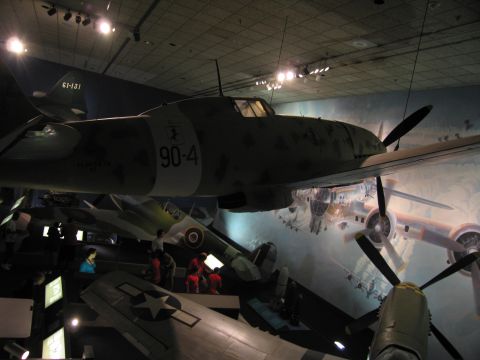 1991 Ford LTD Crown Victoria - twin-engine WWII aircraft coded 90-4 suspended in a museum, with other aircraft and a battle mural visible below.