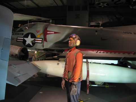 1991 Ford LTD Crown Victoria - mannequin in an orange flight-deck vest and helmet stands before a Navy jet and white missile in a museum.