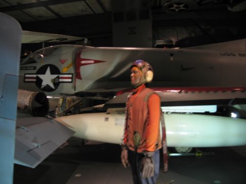 1991 Ford LTD Crown Victoria - flight-deck crewman mannequin in orange gear stands beside a gray Navy jet and ordnance in a museum display.