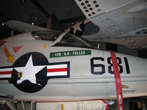 1991 Ford LTD Crown Victoria - gray Navy jet numbered 681 displays a star-and-bars insignia, CDR R.B. Fuller marking and folded wing in a museum.