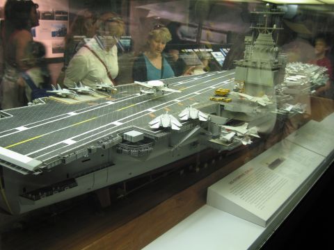 1991 Ford LTD Crown Victoria - Museum visitors look at a large aircraft carrier scale model crowded with deck aircraft inside a glass case.