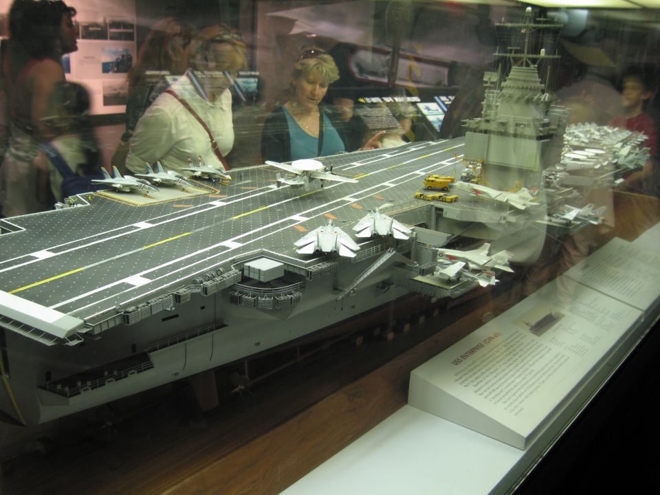 Visitors view a large glass-cased aircraft carrier scale model with deck jets