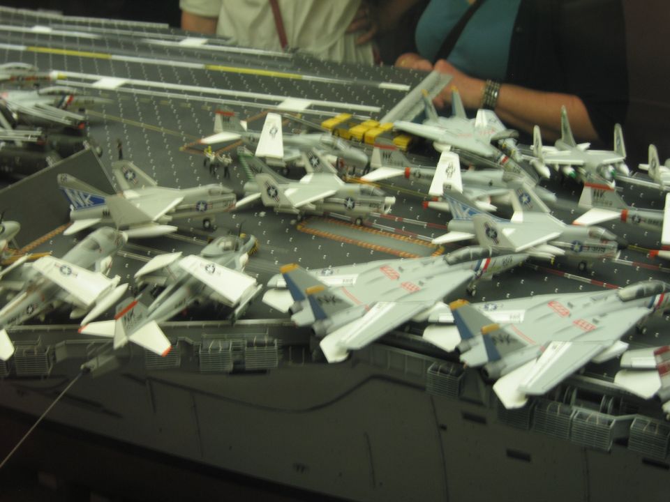 Scale model carrier deck filled with gray Navy fighter jets, people viewing behind