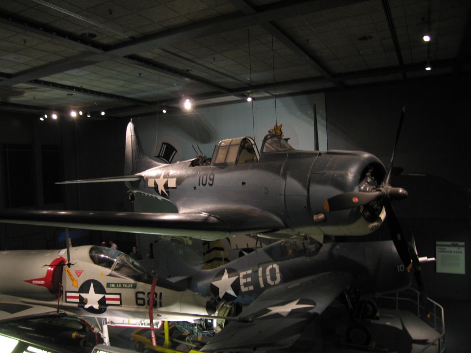 Gray WWII Navy dive bomber 109 with folded wing displayed above other aircraft