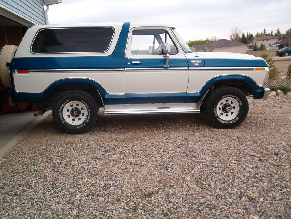 Side view of a blue and white 1978 Ford Bronco with white steel wheels on a gravel driveway
