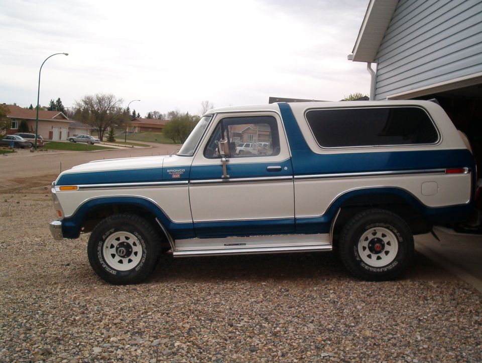 Side view of a two-tone blue and white 1978 Ford Bronco on gravel next to a garage
