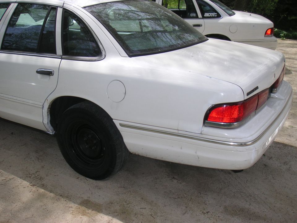 White Ford sedan rear three-quarter on dirt lot with black steel wheels and patched wheel arch