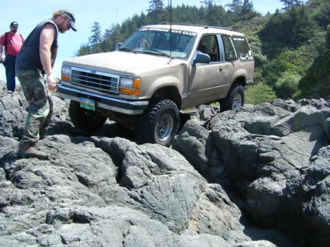 2005 Jeep Wrangler - A tan Ford Explorer is shown off-roading over large rocks, with a person observing its progress.