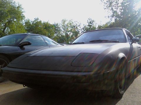 1983 Mazda RX-7 - A dark car with pop-up headlights is parked outside, with another vehicle behind it.