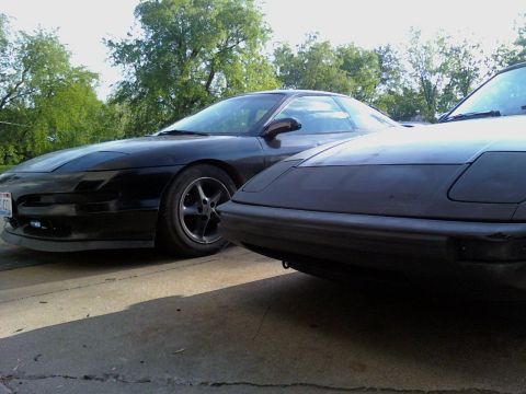 1983 Mazda RX-7 - Two black coupes are parked next to each other on a paved surface with trees in the background.