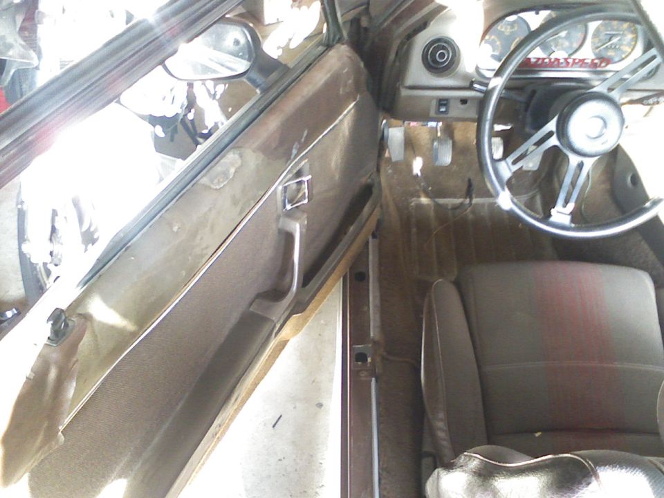 Driver's side interior of a Mazda RX-7 with brown door panel, striped seat, and Mazdaspeed steering wheel area
