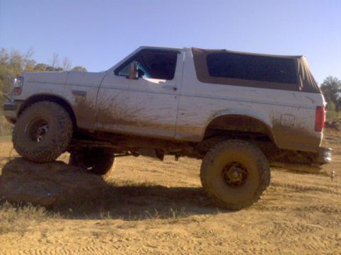 1996 Ford Bronco - Muddy 1996 Ford Bronco with a soft top is shown off-roading over a large rock.