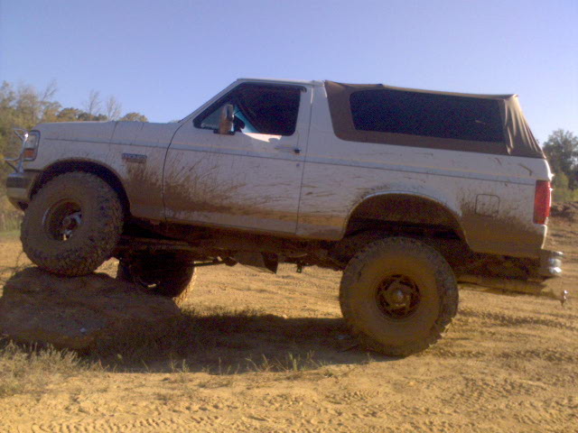 White 1996 Ford Bronco with soft top, covered in mud, driving over a rock
