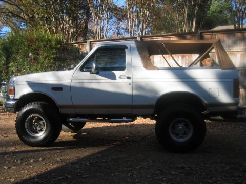 1996 Ford Bronco - Side view of a white lifted Ford Bronco with a tan soft top folded open over the exposed roll bar. It rides on mud-terrain tires with chrom…