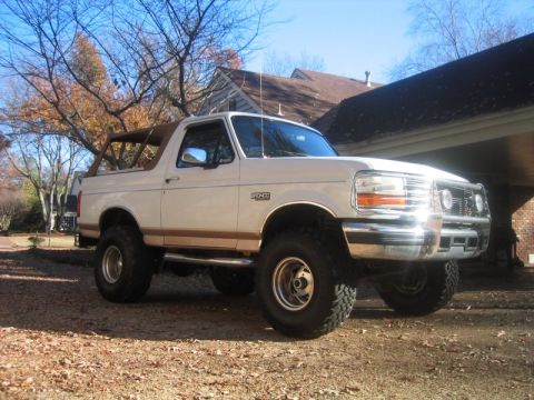 1996 Ford Bronco - Lifted white-over-tan Ford Bronco full-size SUV shot from the front three-quarter angle in a leaf-covered gravel driveway beside a brick ho…