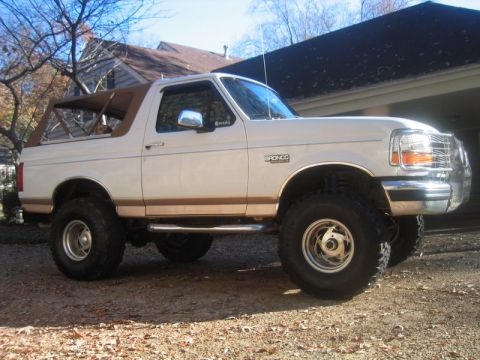 1996 Ford Bronco - Driver-side profile of a lifted white and tan two-tone Ford Bronco parked on a leaf-covered gravel driveway, with the tan top in place and…