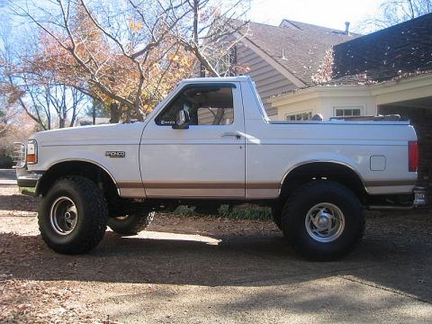 1996 Ford Bronco - Driver-side profile of a white and tan two-tone Ford Bronco with the hardtop removed, lifted on chrome five-slot wheels and oversized mud-t…