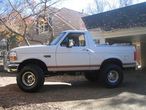1996 Ford Bronco - White lifted full-size Ford Bronco with tan trim and chrome wheels parked on a driveway by a brick house in autumn.