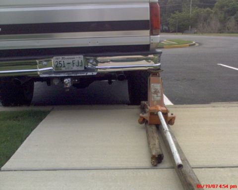 1996 Ford Bronco - Rear view of a white and tan two-tone SUV being raised by an orange floor jack placed under the passenger side of the chrome rear bumper, w…