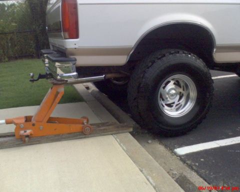 1996 Ford Bronco - Rear corner of a white and tan two-tone full-size SUV lifted with an orange floor jack under the chrome rear bumper, parked over a wood pla…