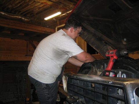 1979 Ford Bronco - A man in a light-colored t-shirt and dark pants works on the engine bay of a vehicle with the hood open. A red cordless drill rests on the…