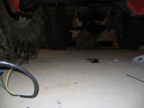 1979 Ford Bronco - A person is working underneath the chassis of a large vehicle, likely a truck or SUV, with oversized tires visible. Wires and a dark fluid…