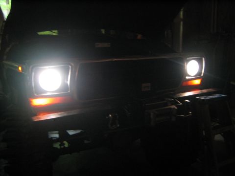 1979 Ford Bronco - The front of a dark-colored vehicle, likely a 1979 Ford Bronco, is illuminated by its bright headlights and orange turn signals. A winch is…