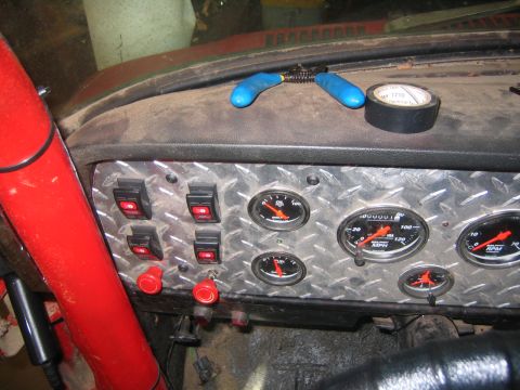 1979 Ford Bronco - A close-up view of a custom dashboard in a vehicle, featuring a diamond plate panel with multiple gauges and toggle switches. A red steerin…
