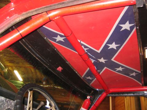 1979 Ford Bronco - The interior of a vehicle shows a red roll cage and a panel painted with a Confederate flag design on the ceiling. A fire extinguisher is v…