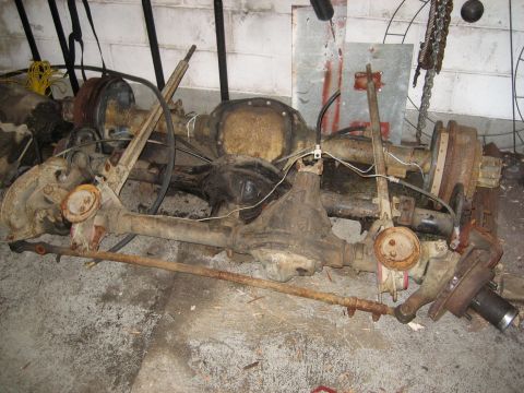 1990 Ford Bronco - A pile of rusty Ford Bronco front and rear axles with brake components and steering linkages are laid out on a concrete floor. The parts ap…