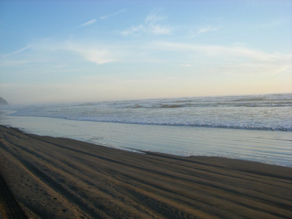 Sandy beach with tire tracks leading into the ocean under a blue sky.