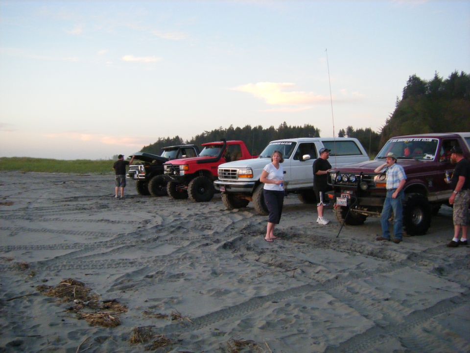 Group of lifted Ford Bronco SUVs parked on a sandy beach with people standing around them.