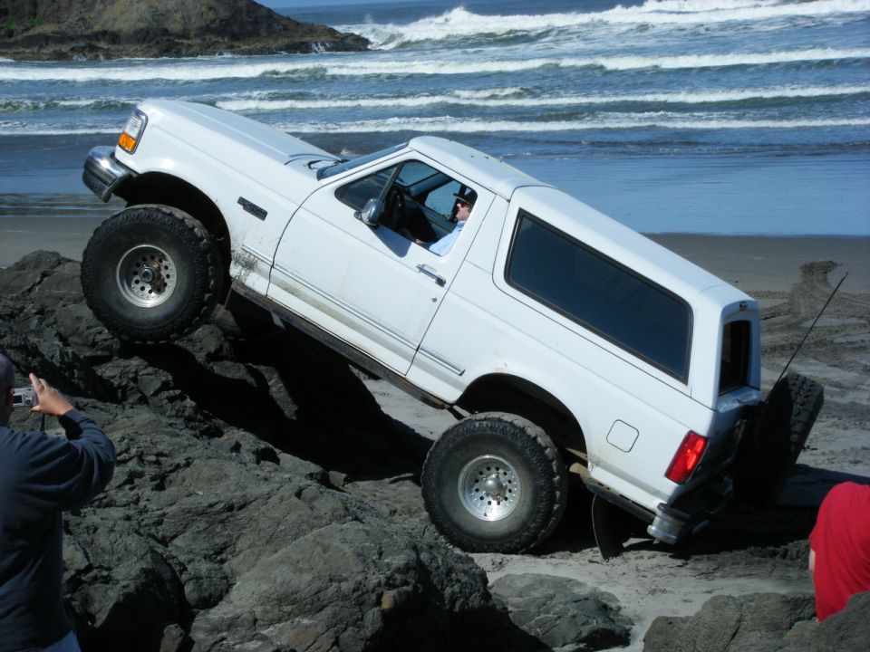 White 1990s Ford SUV climbing over rocks on a beach.