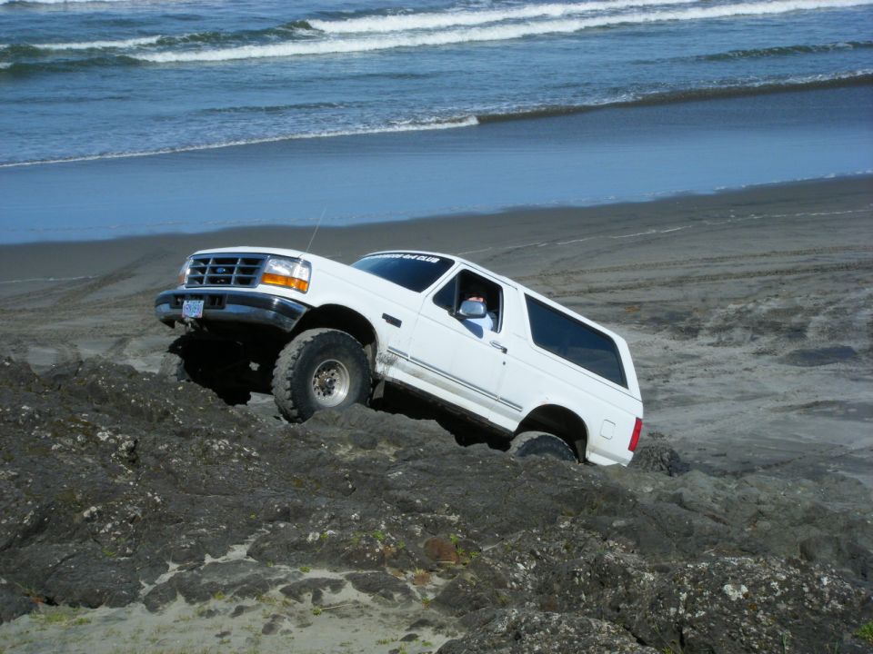 White Ford Bronco driving on a rocky beach near the ocean.