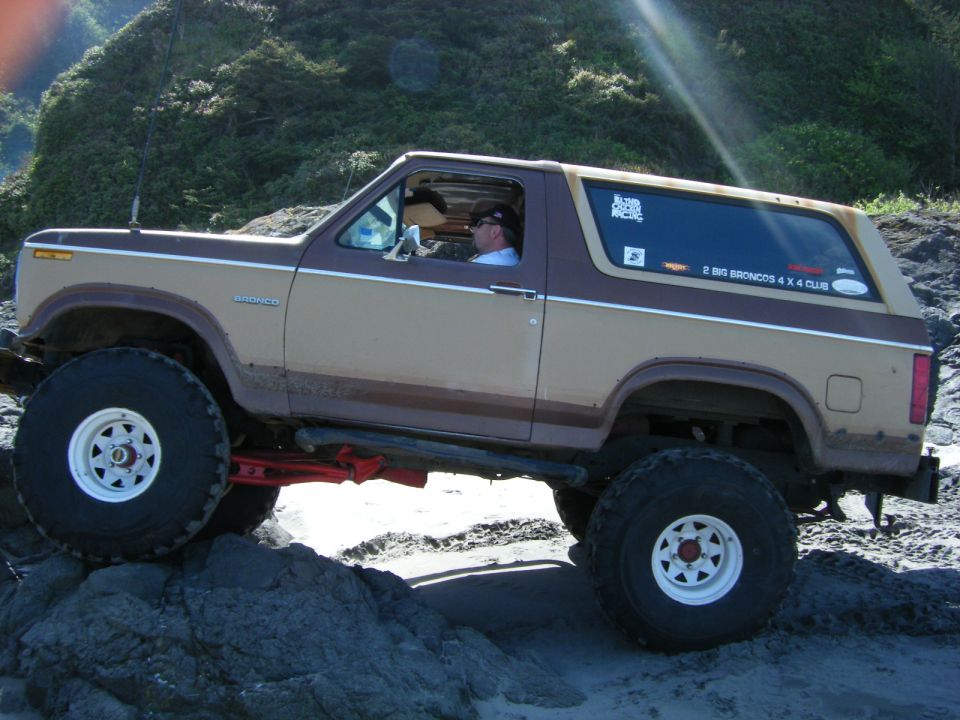 Lifted two-tone brown and tan Ford Bronco driving over rocks on a beach.