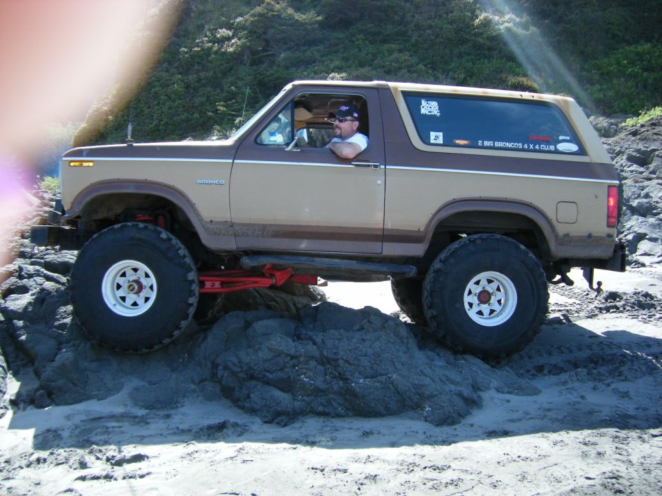Lifted two-tone brown and tan Ford Bronco off-roading on rocks near a beach.