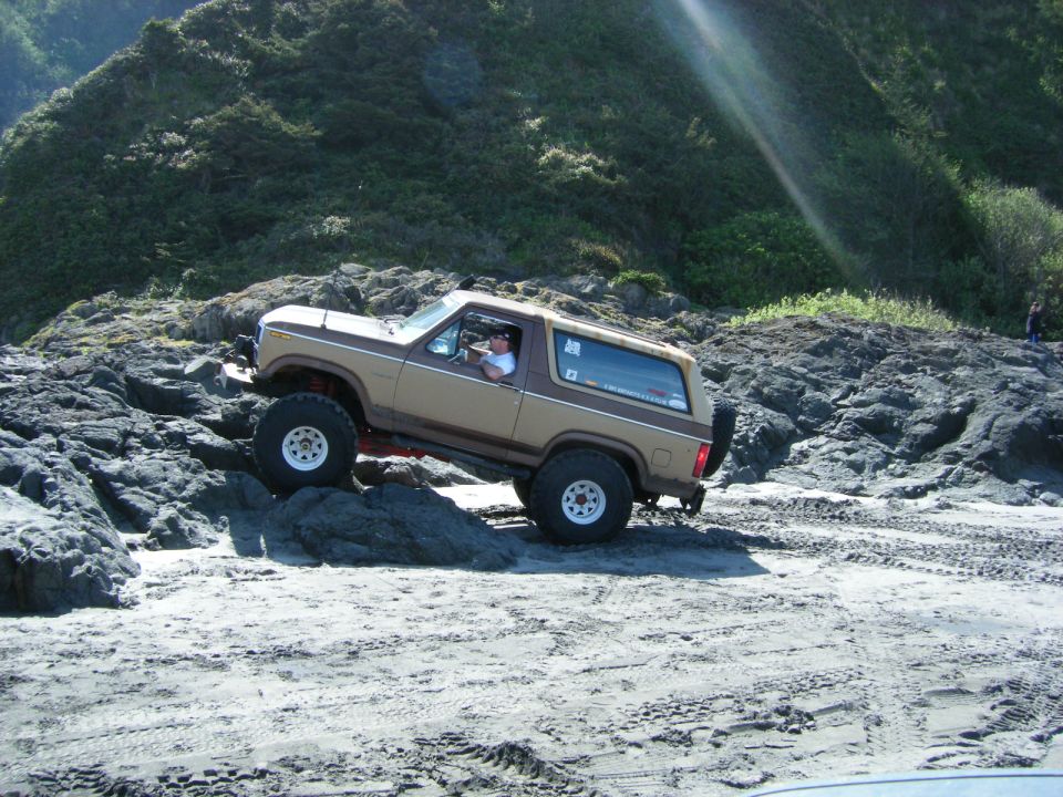 Tan and brown Ford Bronco driving over rocks on a beach.