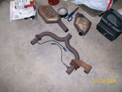 1995 Mercury Grand Marquis - Removed exhaust hardware laid out on a garage floor: a rusty Y-pipe with flanged ends and an attached oxygen-sensor wire, plus two cut-open…