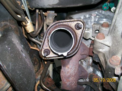 1995 Mercury Grand Marquis - A close-up view of a rusted exhaust manifold flange and pipe connection on a vehicle engine. The flange shows signs of wear and a broken ga…