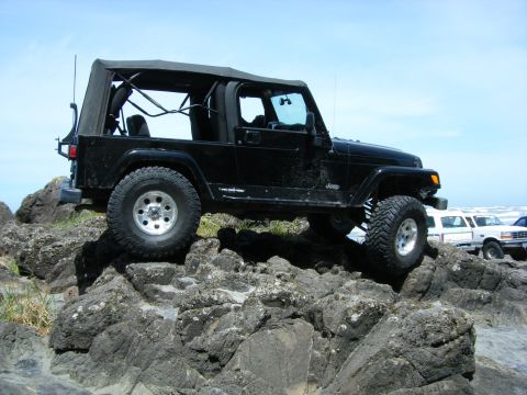 1994 Ford Explorer - A black Jeep Wrangler TJ navigates rocky terrain near the ocean, with other vehicles parked on the beach in the background.