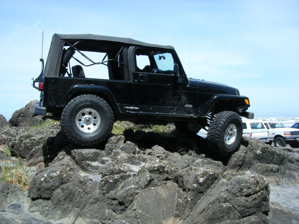 Black Jeep Wrangler TJ on rocks, side view. Other vehicles visible in background