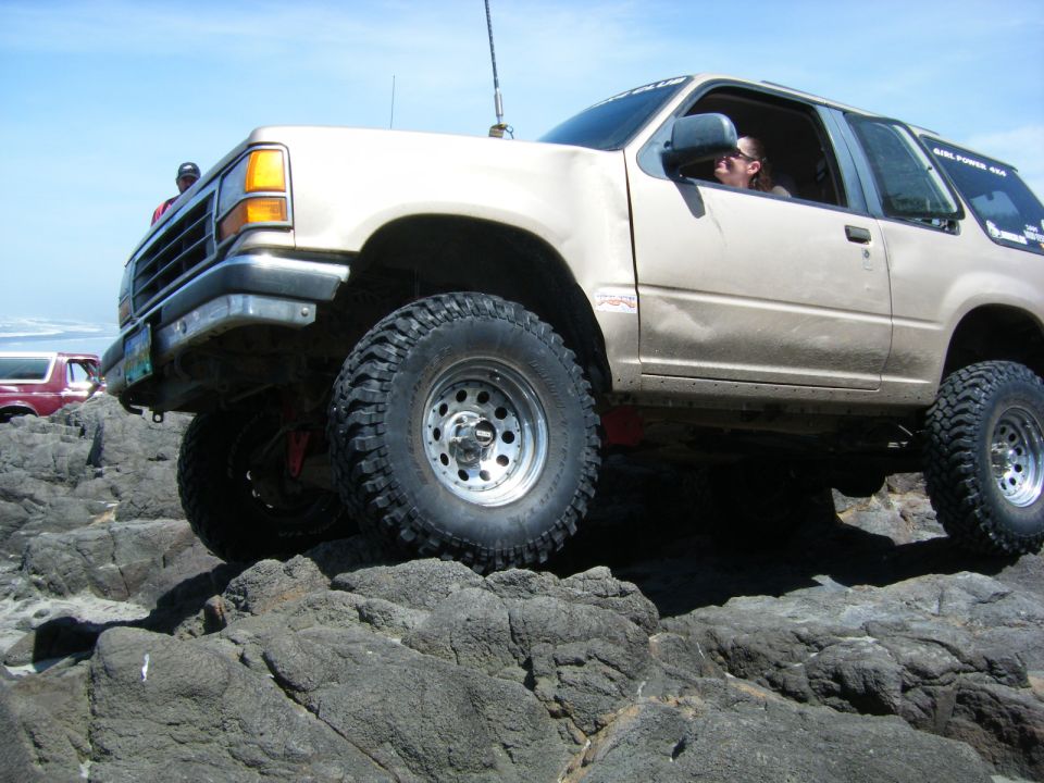 Lifted tan 1990s Ford Explorer off-roading over rocks on a beach.