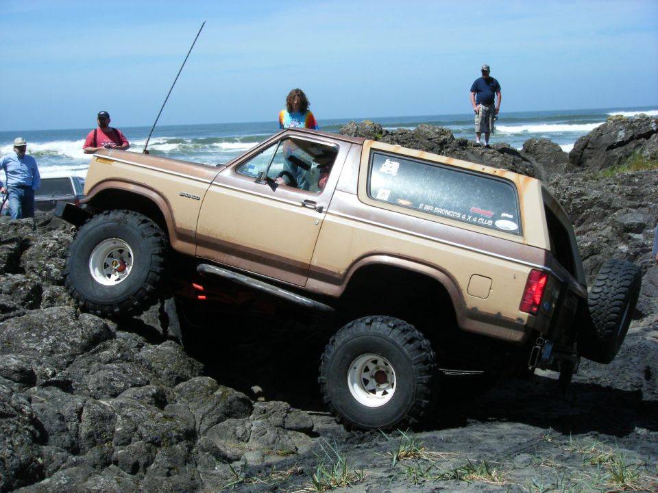 Tan and brown Ford Bronco on a rocky beach with large tires, people nearby.