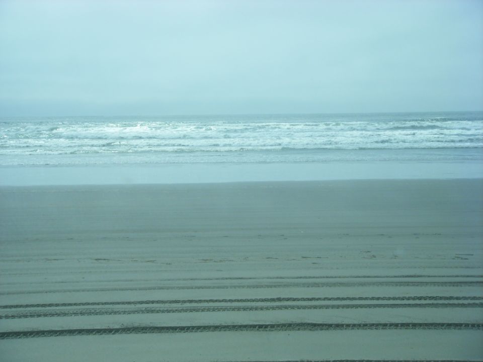 Sandy beach with tire tracks and ocean waves under a hazy sky.