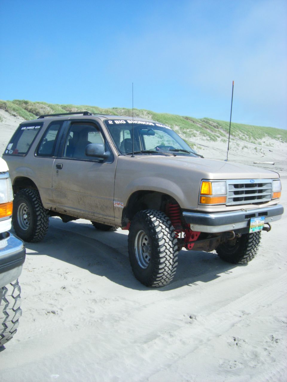 Tan 1994 Ford Explorer with large off-road tires parked on a sandy beach next to another vehicle