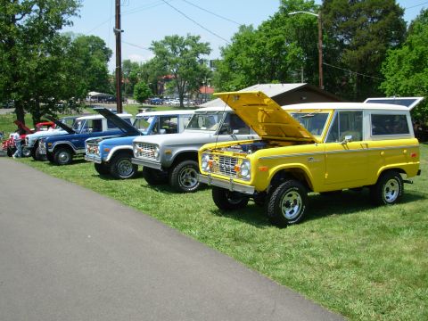 1983 Ford Bronco - A row of Ford Broncos, including a yellow one with its hood up, are displayed on a grassy field under a sunny sky.